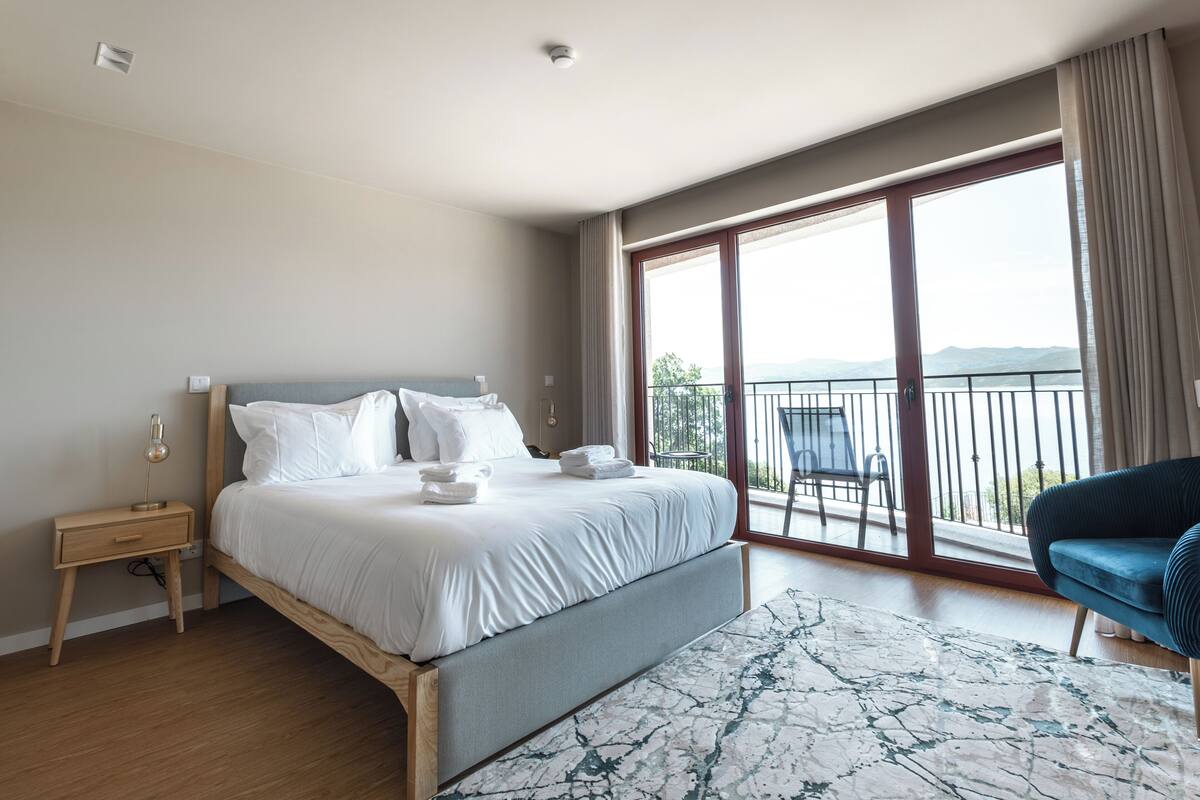 A modern hotel room with a large bed, a bedside table, a chair, and a balcony with a view.