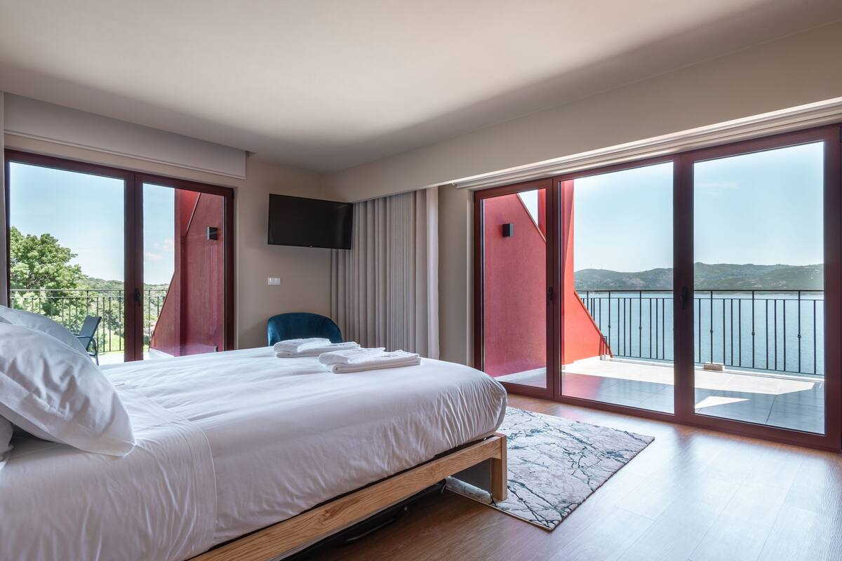 A modern bedroom with a large bed, a TV mounted on the wall, and a balcony with a view of the sea.