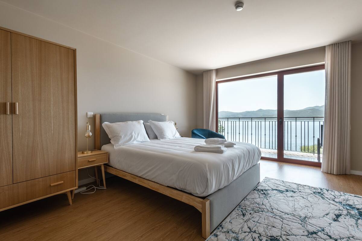 A modern bedroom with a large bed, wooden wardrobe, and a view of the sea.