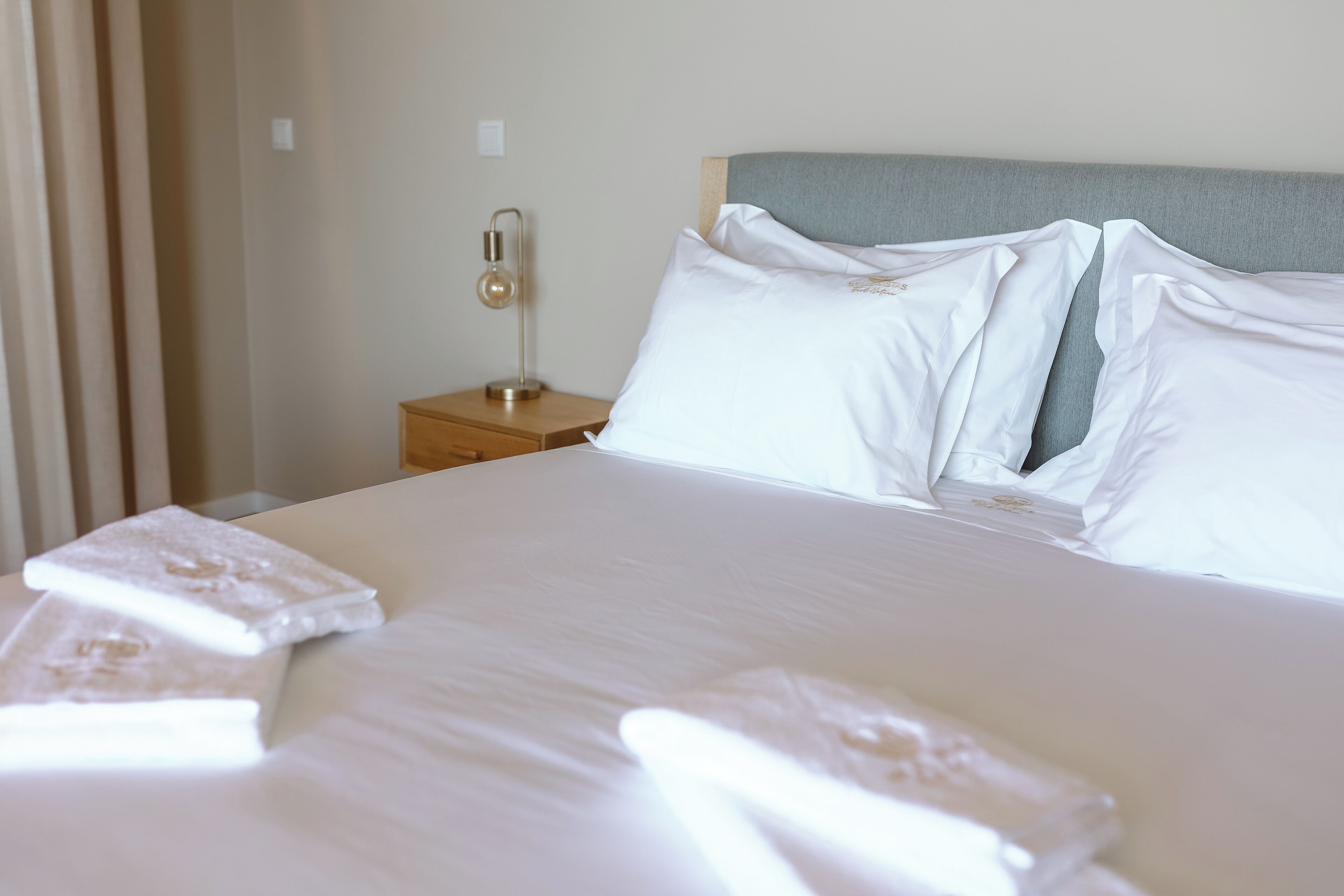 Una cama bien hecha con sábanas y almohadas blancas, una mesita de noche con lámpara y un cabecero gris.