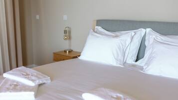 Una cama bien hecha con sábanas y almohadas blancas, una mesita de noche con lámpara y un cabecero gris.