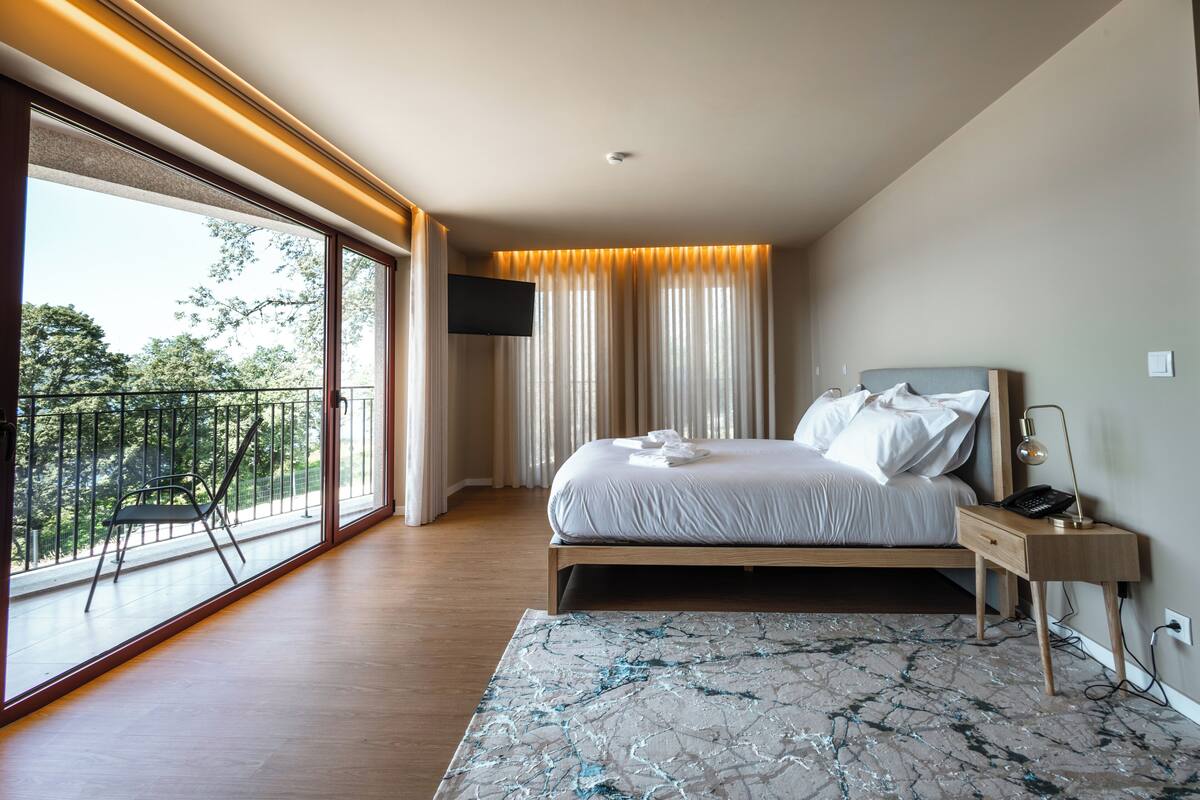 A modern bedroom with a large window, a bed with white linens, a desk with a lamp, and a view of greenery outside.