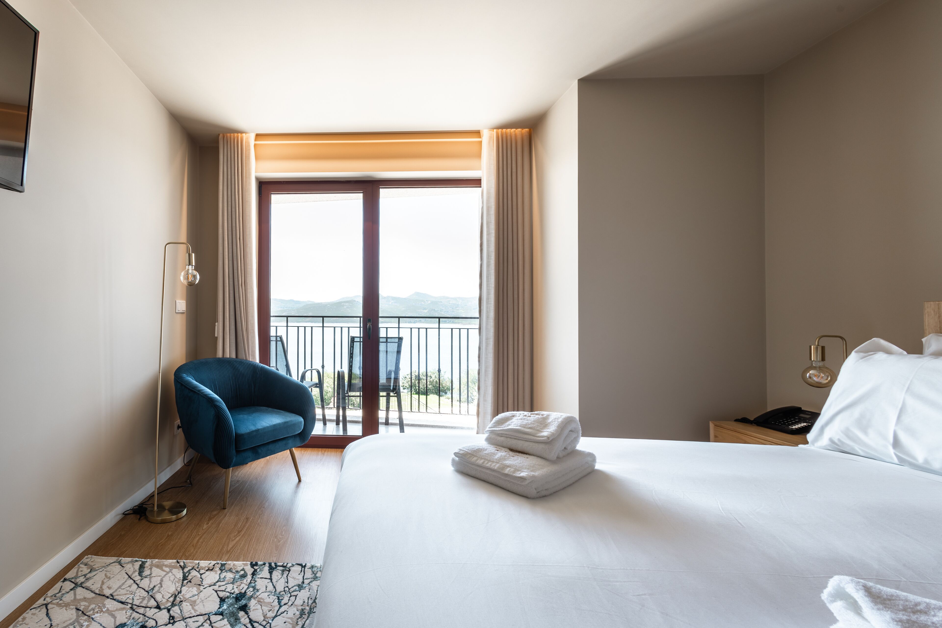 Una habitació d'hotel amb un llit gran, una cadira blava, un balcó amb vistes i una taula de treball amb una làmpada.