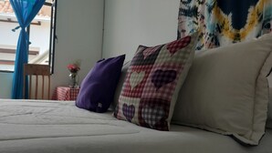 A bed with a checkered pillow and a plain pillow, a vase with flowers, and a window with blue curtains. - Family Host (Villa de Leyva)