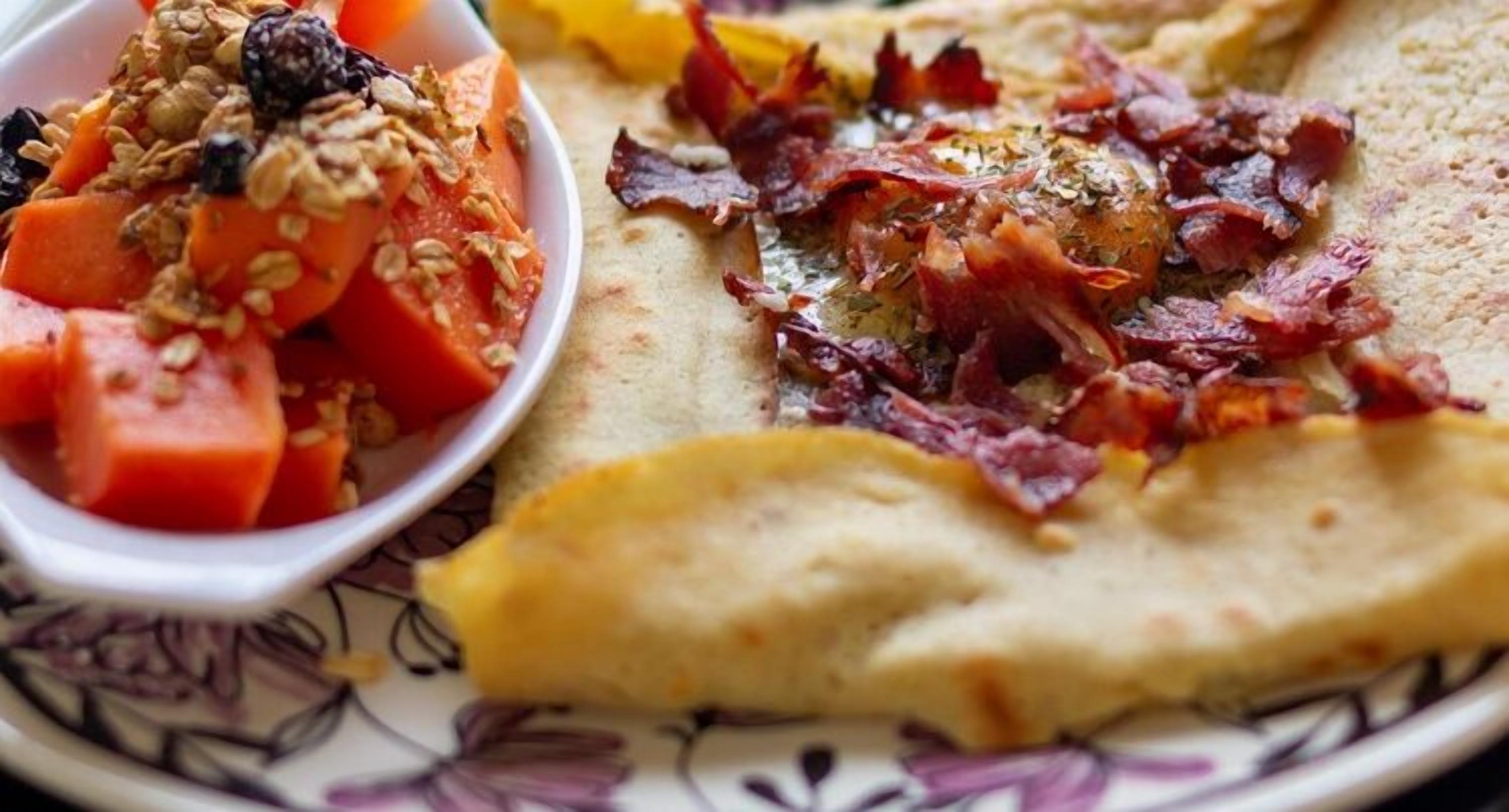 En tallrik med ett plattbröd täckt med bacon och en skål med fruktsallad och granola.