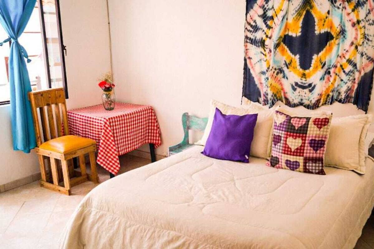 A room with a bed, a small table with a checkered tablecloth, a chair, and a colorful tapestry on the wall.