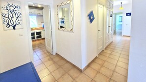A hallway with white walls, a tiled floor, and a door leading to a kitchen. There are wall-mounted light fixtures and a mirror. - Casa Kody - with BBQ, Fiber optic, private parking and silence.  (La Oliva)