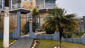 Une maison à deux étages avec une façade en pierre, un toit bleu et une cour clôturée où se trouve un palmier.