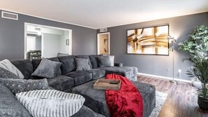 A living room with a sectional sofa, a red throw blanket, and a large abstract painting on the wall. - Hoff Gardens 9 - 2BR 2BA - Downtown Lubbock (Lubbock)
