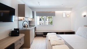 Ein modernes Hotelzimmer mit Bett, einer kleinen Küchenzeile, einem Fernseher und einem Fenster mit Blick auf Gebäude.