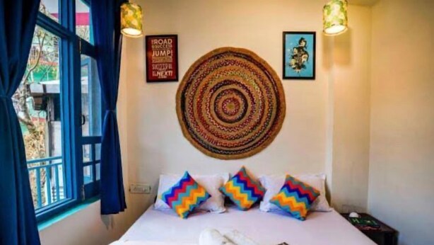 Una habitación de hotel con una cama, almohadones con diseño en zigzag y colores vivos, un tapiz de pared tejido y un cartel motivacional.
