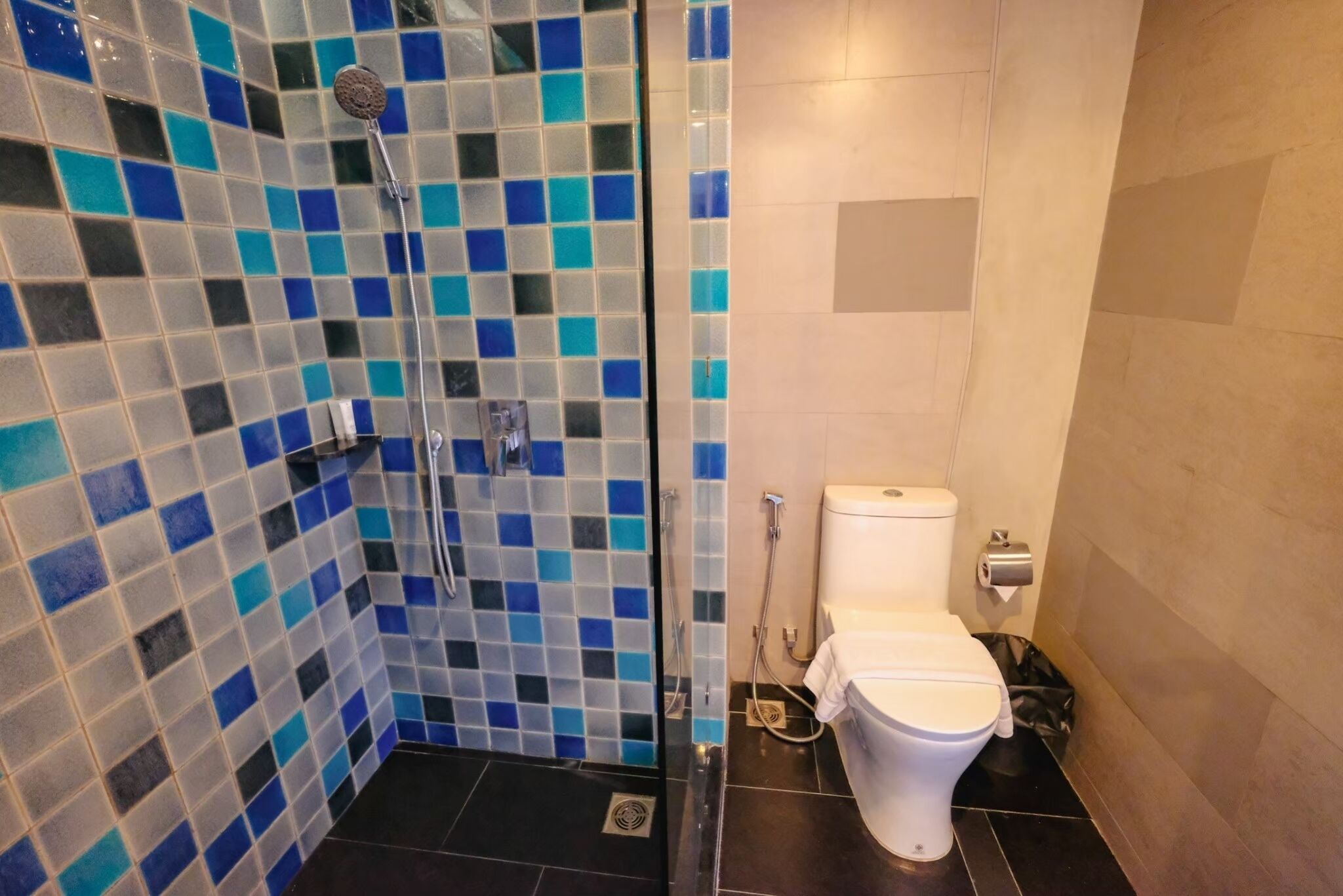 A bathroom with a shower area featuring blue and white mosaic tiles, a white toilet, and a black trash bin.