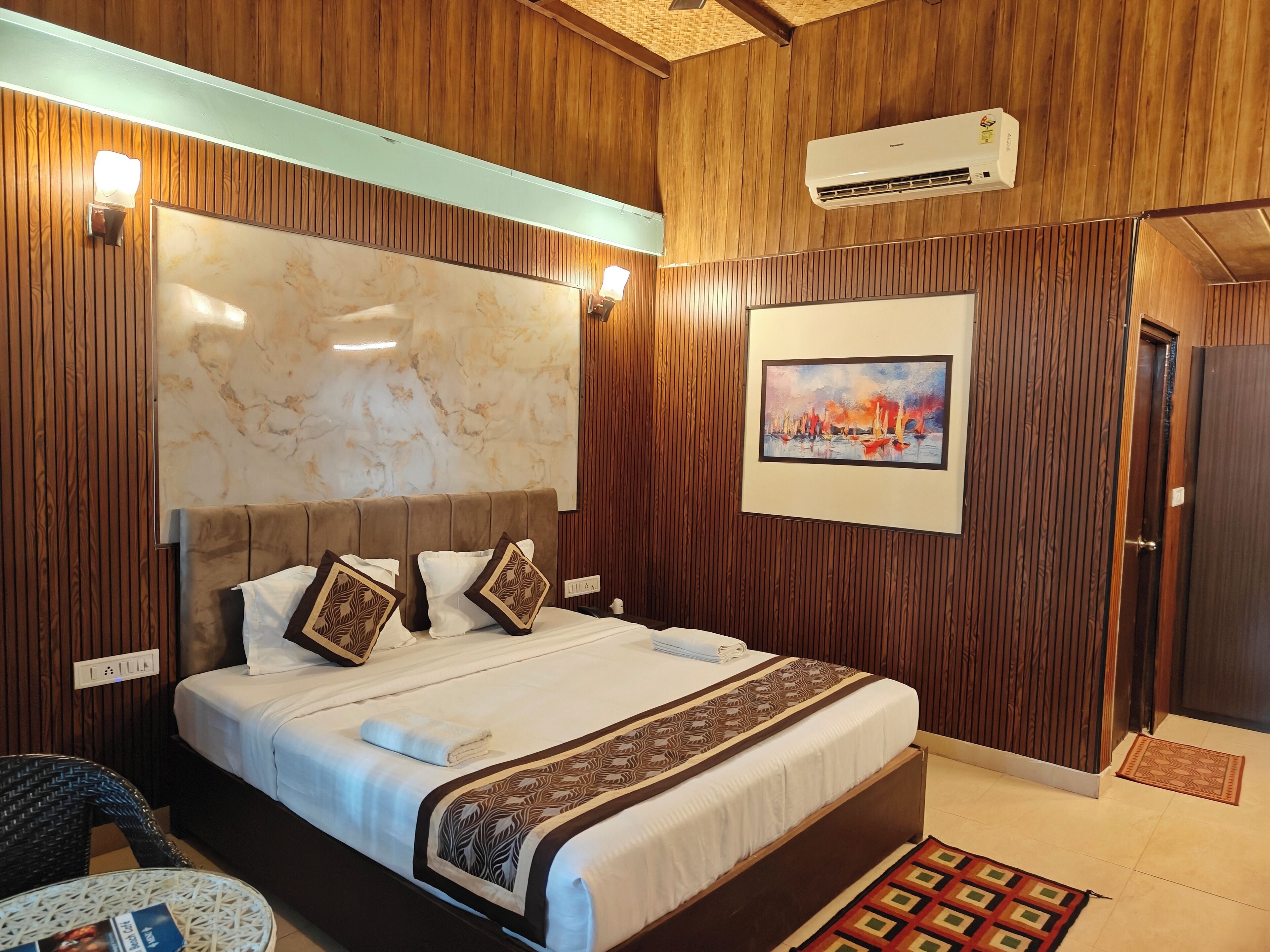 A hotel room with a wooden paneled wall, a marble headboard, a bed with white bedding and patterned pillows, a painting on the wall, and an air conditioning unit.