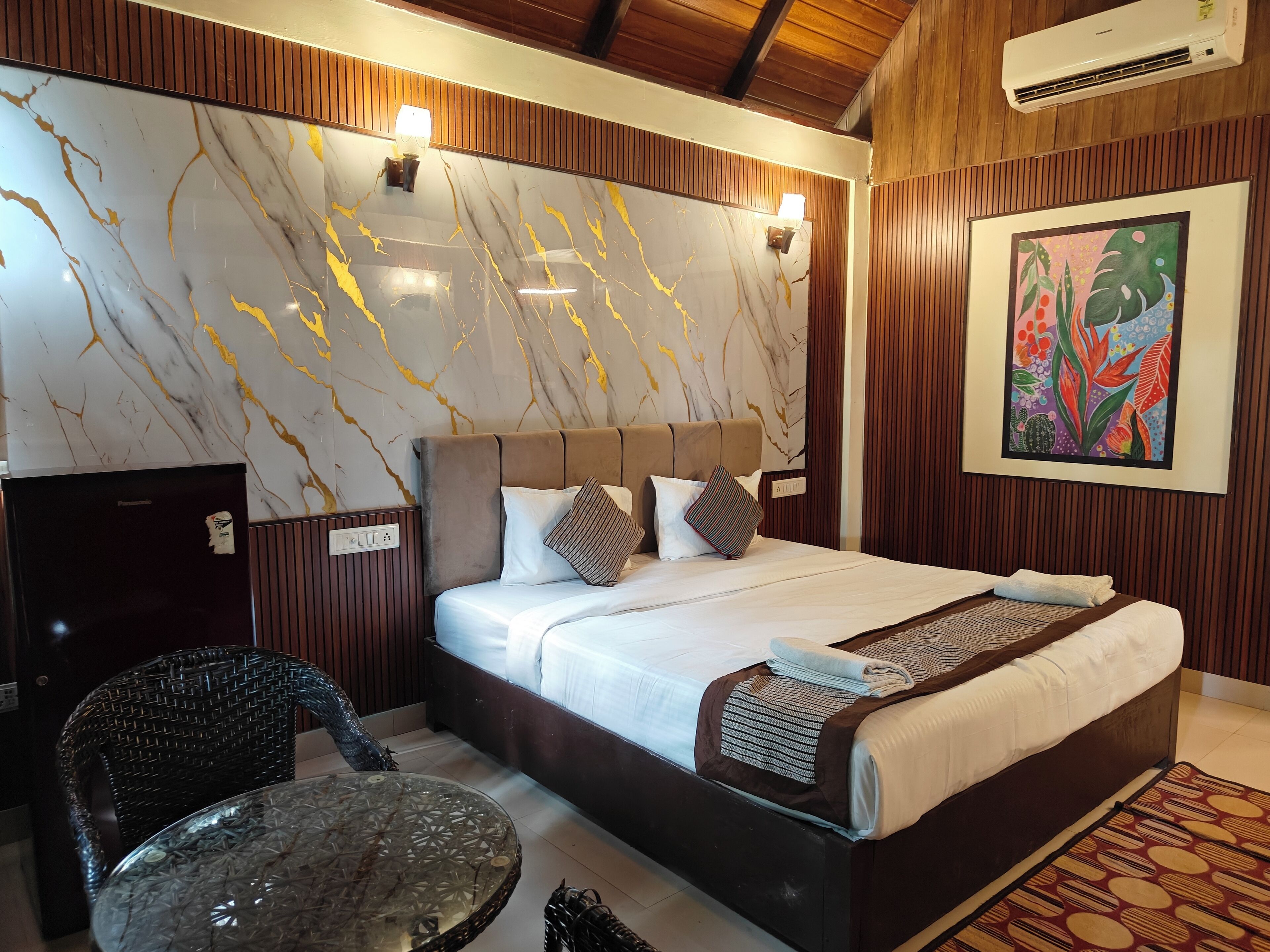 A neatly arranged hotel room with a marble-patterned wall, a bed with a brown headboard, and a colorful artwork on the wall.