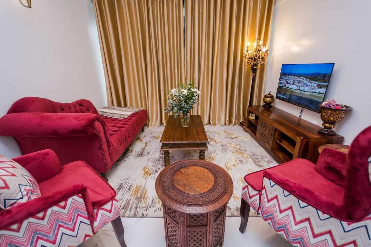 A living room with red velvet sofas, a wooden coffee table, a patterned rug, and a television displaying a scenic view.