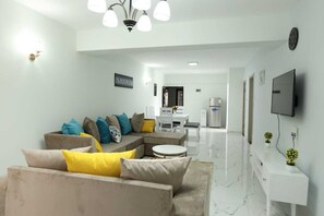 A modern living room with a sectional sofa, a round coffee table, and a TV mounted on the wall. - Lux Suites Sandalwood Gardens Apartments (Nairobi)