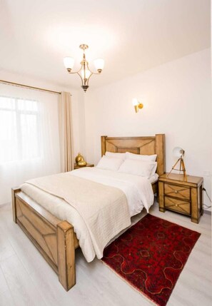 A bedroom with a wooden bed, a red patterned rug, a bedside lamp, and a chandelier. - Lux Suites Sandalwood Gardens Apartments (Nairobi)