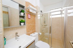 A modern bathroom with a glass-enclosed shower, a white sink, and a toilet. There are shelves with potted plants and a towel rack. - Lux Suites Sandalwood Gardens Apartments (Nairobi)