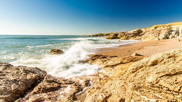 Une plage rocheuse où les vagues se brisent sur le rivage, un ciel dégagé et une vue lointaine sur des falaises.