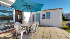 Une terrasse avec une table et des chaises, un parasol bleu et un bâtiment blanc aux volets bleus.