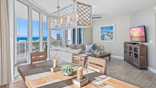 A spacious living room with a large wooden dining table, a sofa, and a TV on a wooden cabinet. The room has large windows with a view of the ocean and buildings.