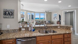 A kitchen with granite countertops, stainless steel sink, and wooden cabinets. There is a large window with a view of the beach and buildings.