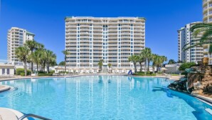 A large swimming pool with a clear blue water, surrounded by palm trees and a tall hotel building in the background.