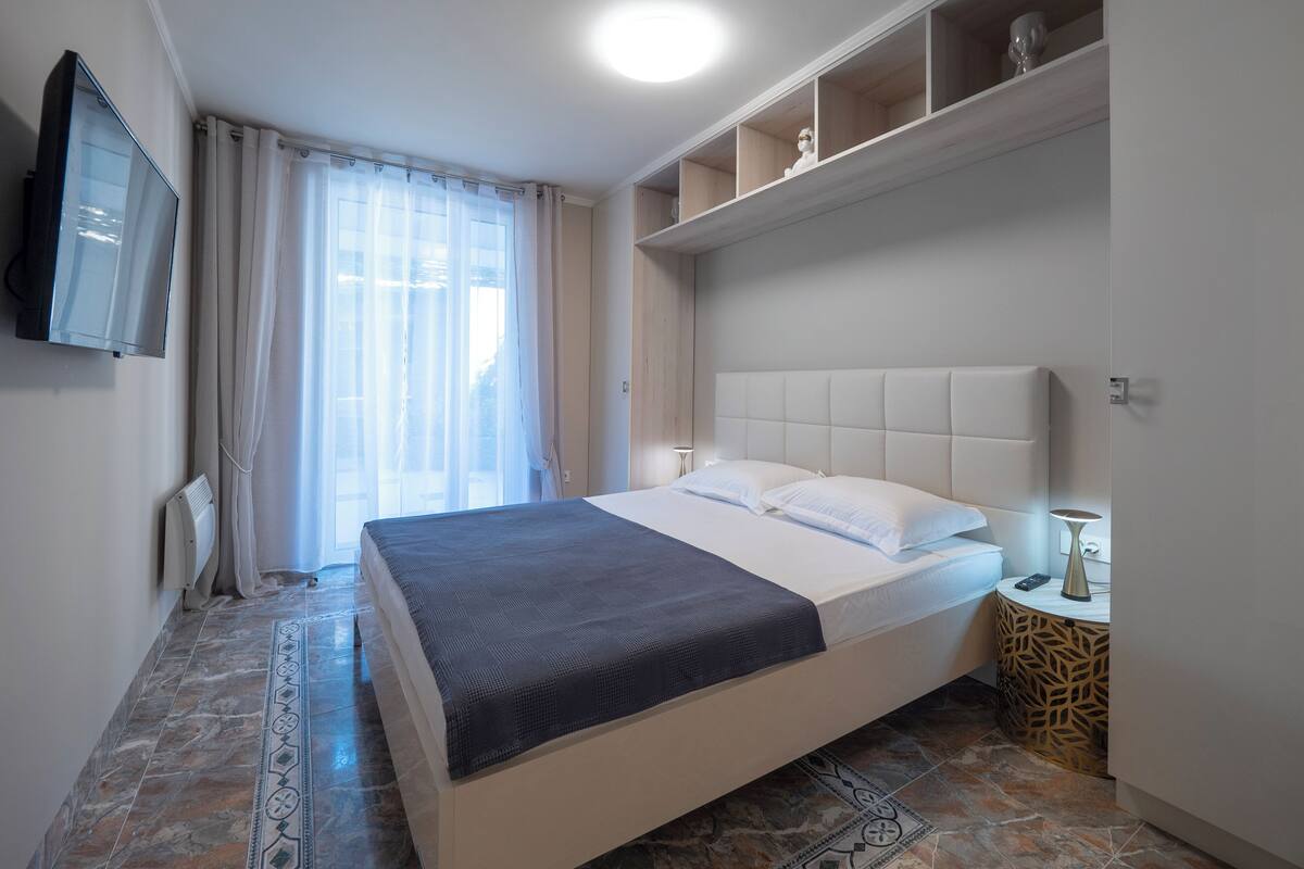 Quarto de hotel moderno com uma cama grande, uma TV de tela plana, uma mesinha auxiliar e uma prateleira decorativa.
