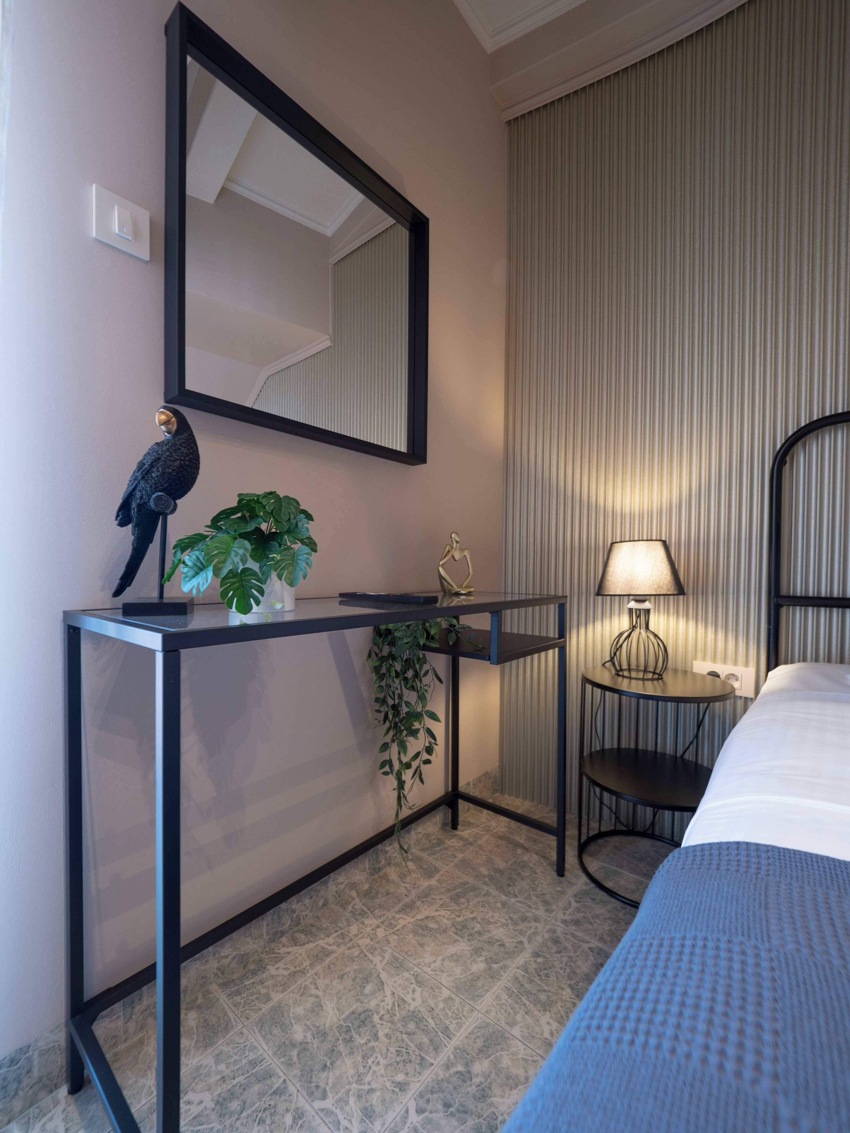 A hotel room with a bed, a bedside table with a lamp, a mirror, a potted plant, and a decorative sculpture.