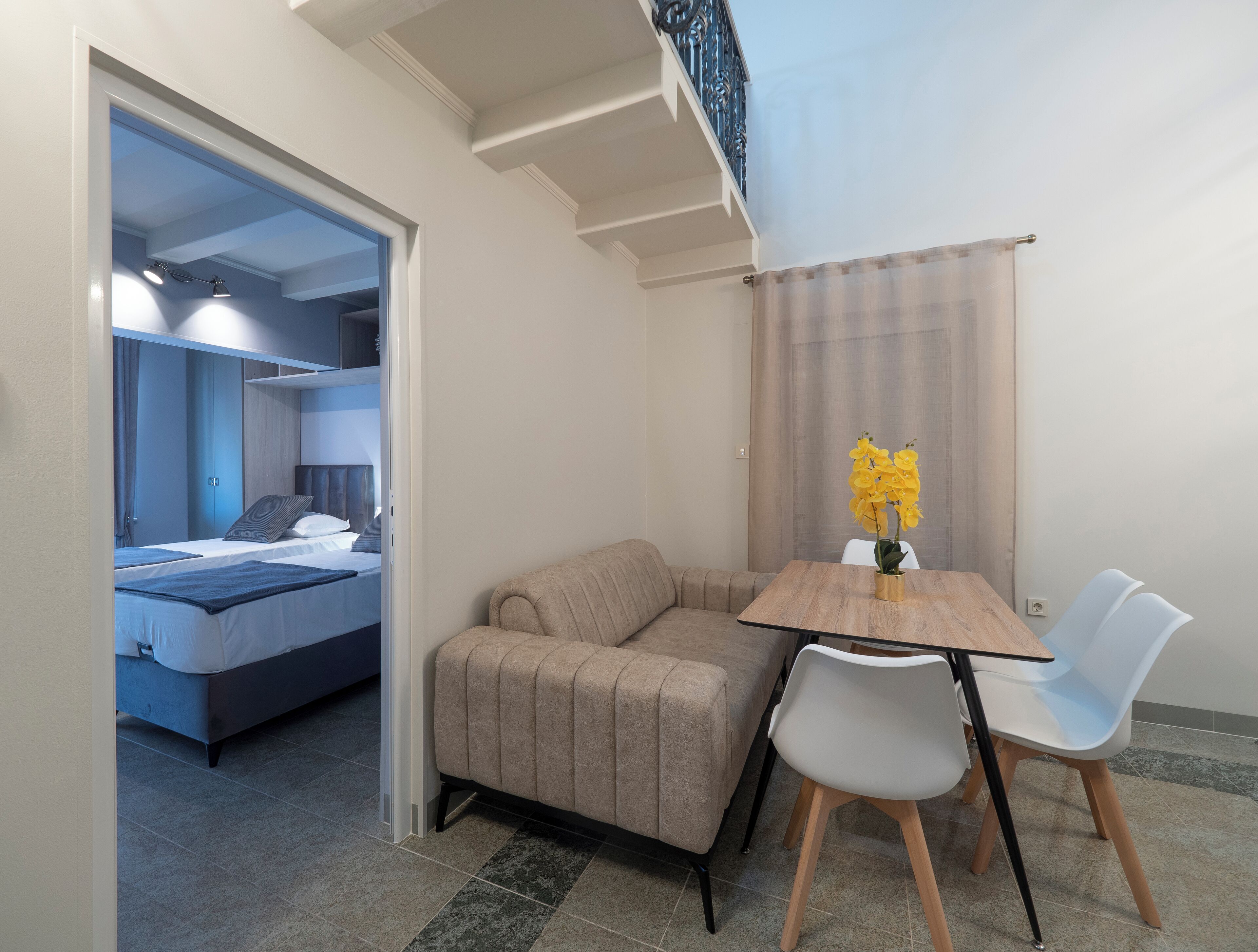 A modern hotel room with a bed, a sofa, a dining table with chairs, and a small table with a vase of flowers.