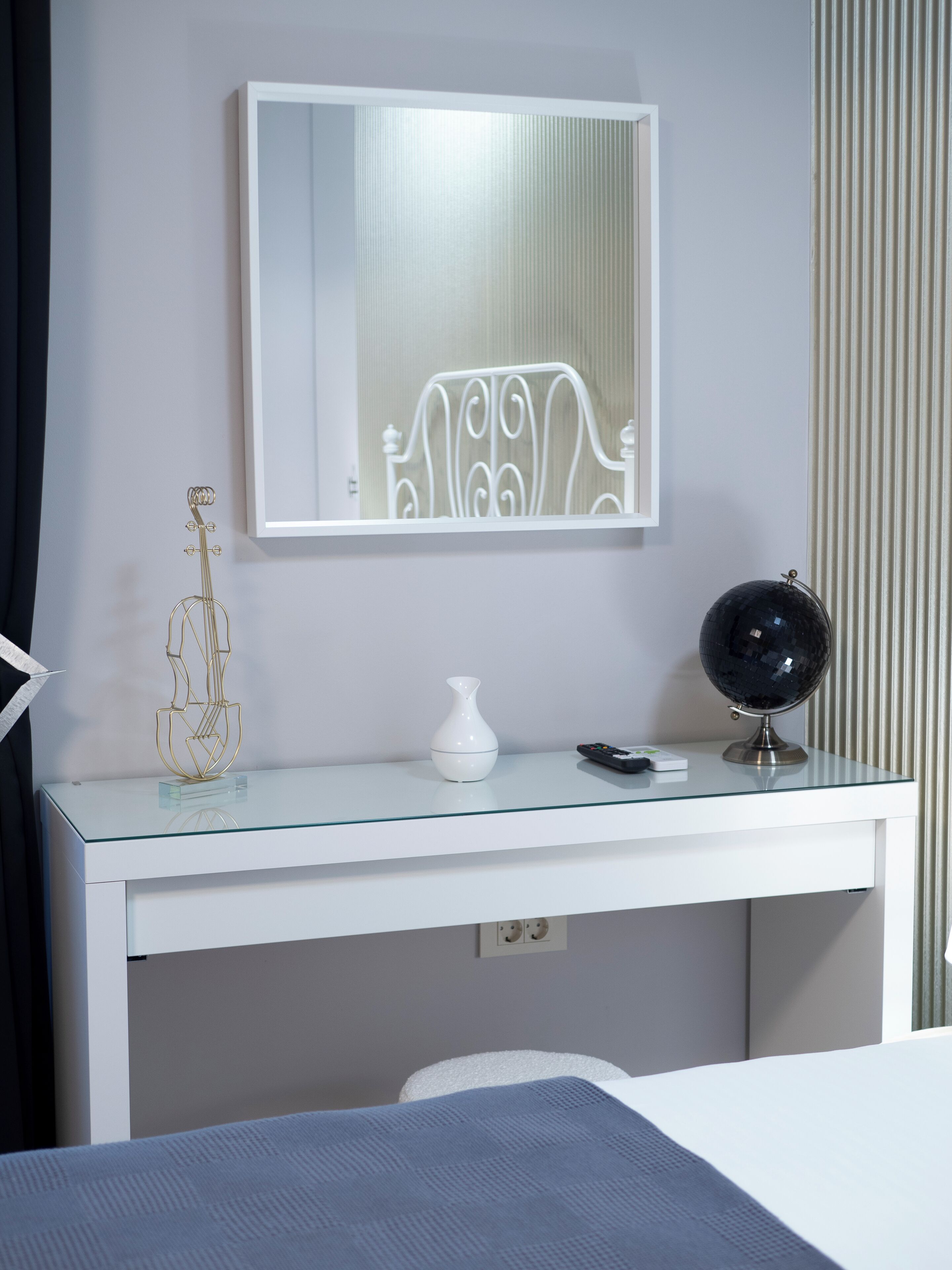 A modern bedroom with a glass desk, a white vase, a black globe, and a framed mirror.