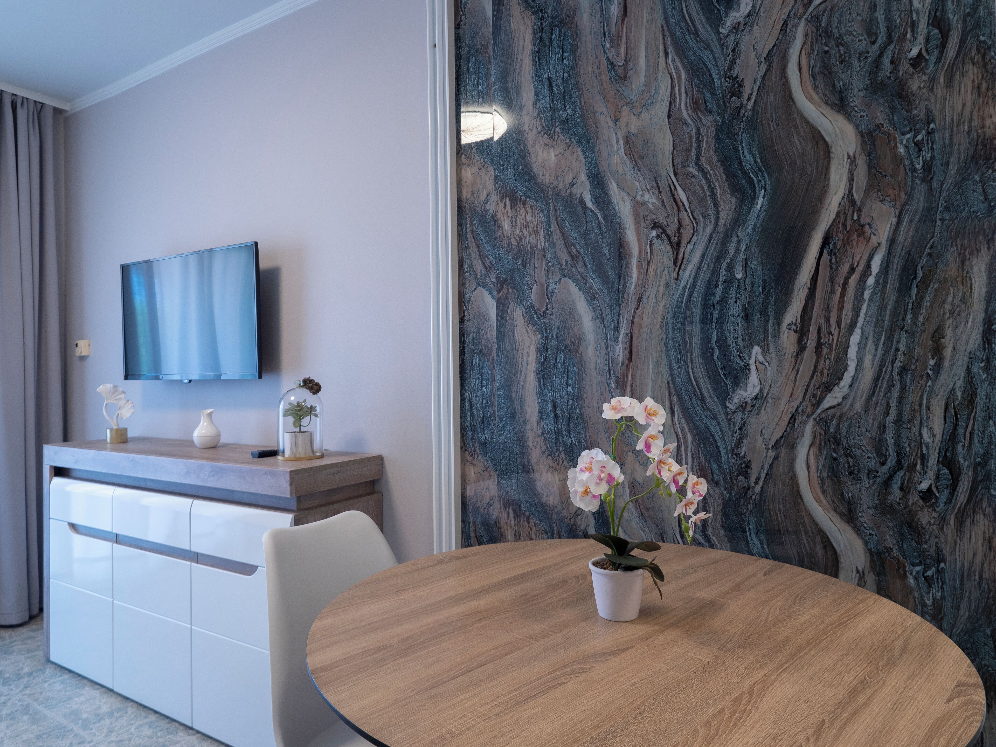 A modern room with a marble wall, a flat-screen TV, a wooden table with a potted orchid, and a white cabinet.