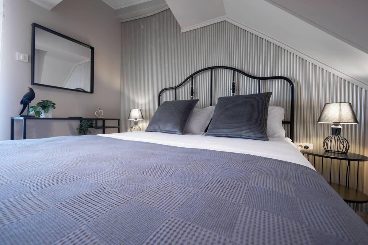 Quarto de hotel moderno com uma cama grande, uma mesinha de cabeceira, um abajur e um espelho fixado na parede.