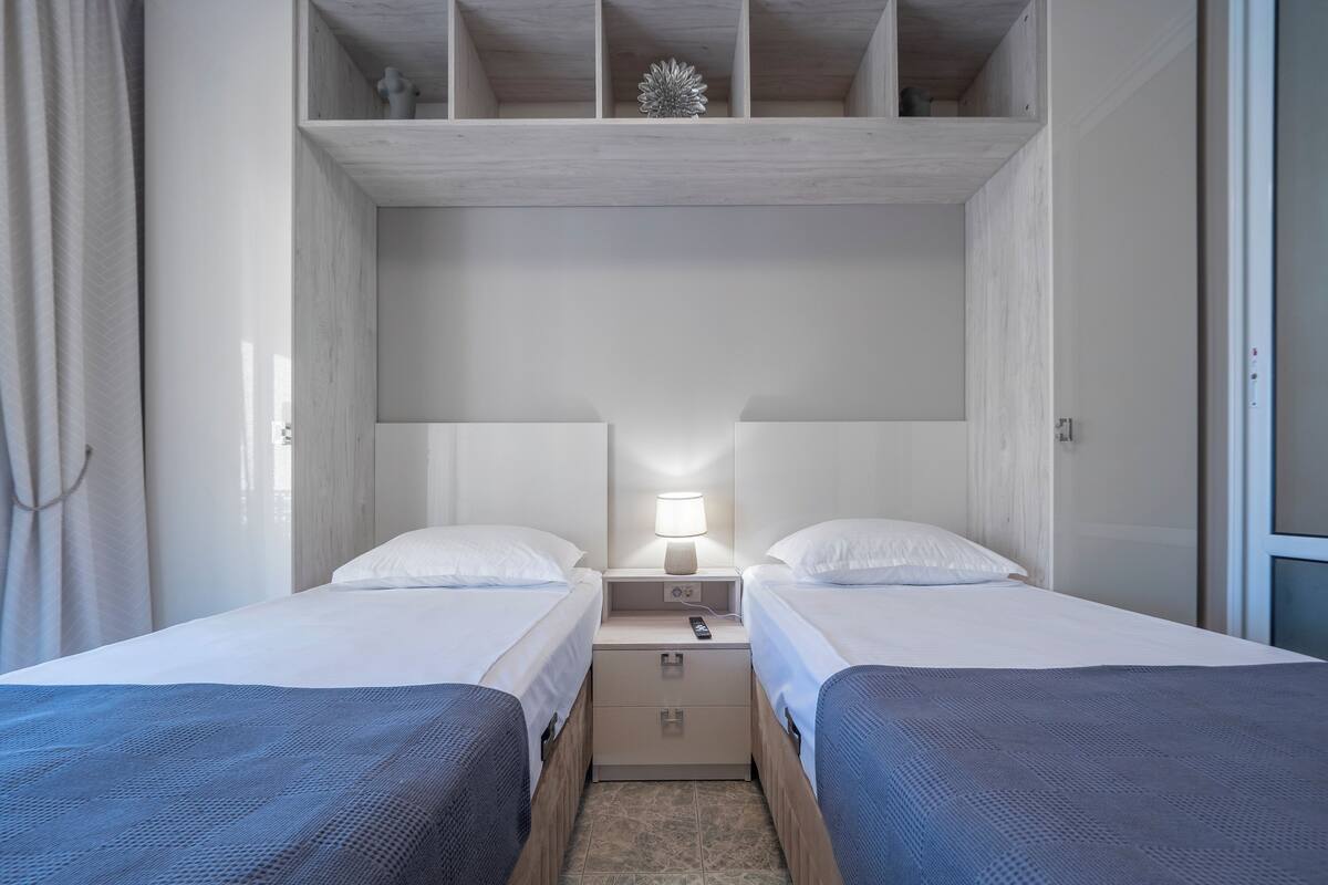Quarto de hotel com duas camas de solteiro, uma mesa de cabeceira com abajur e um pequeno armário.