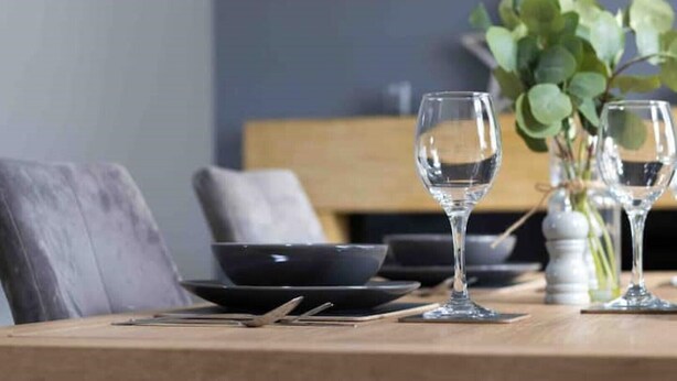 Mesa de jantar com duas taças de vinho, uma tigela e um vaso com folhas verdes.