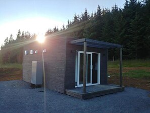 A small, modern building with a glass door and a covered porch, surrounded by a forested area. - Buubble Hotel-Ölvisholt (Selfoss)