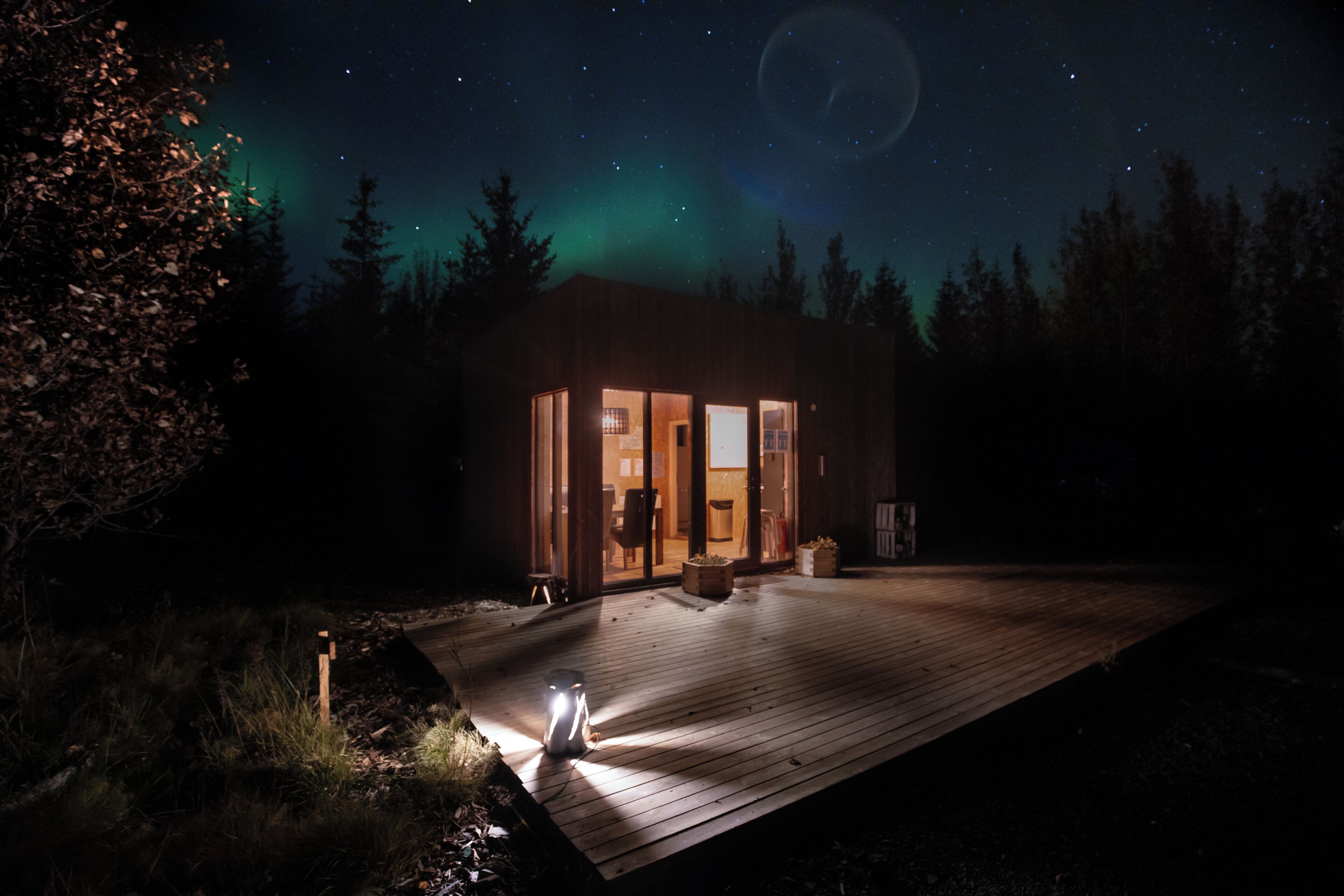 A wooden cabin with glass doors, a deck, and a night sky with stars and the aurora borealis.