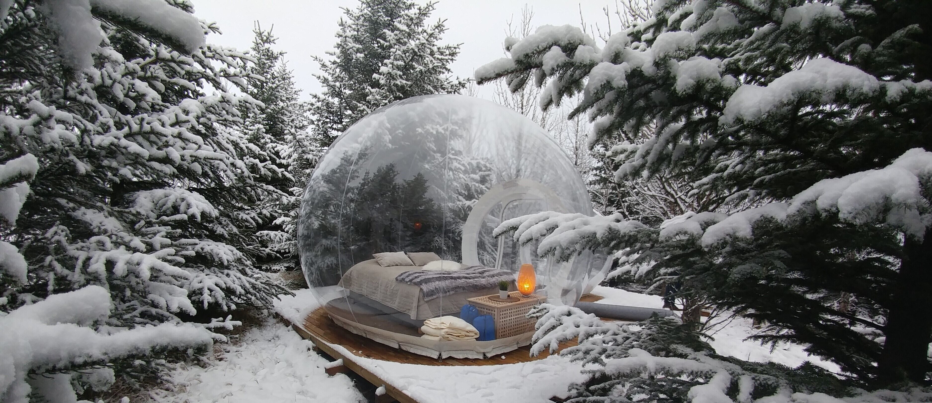 A snow-covered outdoor area with a transparent dome structure containing a bed and a lantern.