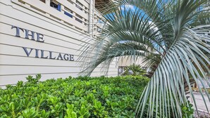 Un edificio con el letrero "THE VILLAGE", rodeado de arbustos verdes exuberantes y una palmera.