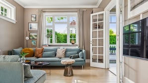 A living room with a sofa, a coffee table, and a side table with a plant. There is a window with curtains, a door leading outside, and a bookshelf. - Beautiful Villa, 150 m2 activity hall with tabletennis etc. outdoor 6 pers. spa (Taastrup)