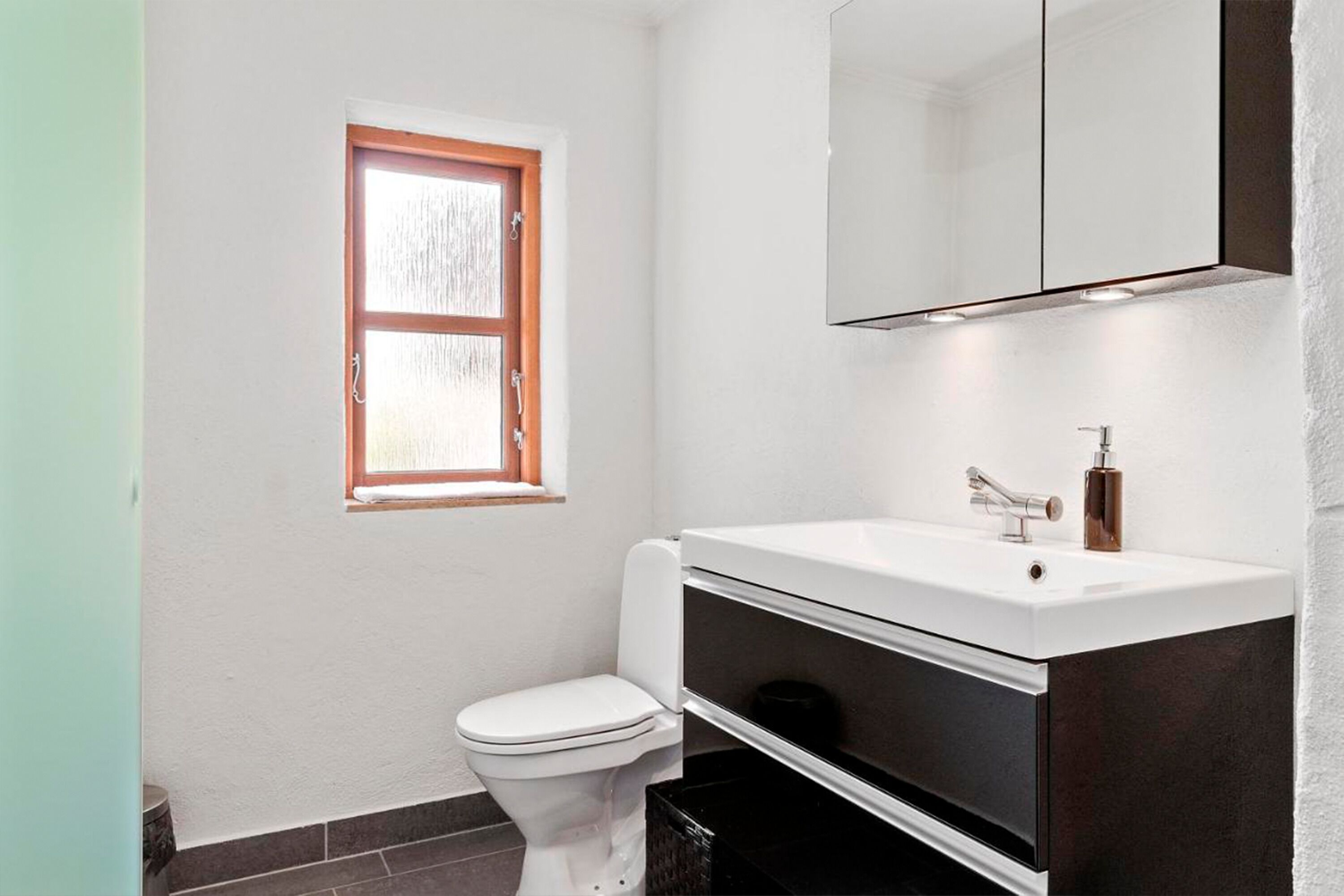 A modern bathroom with a white toilet, a white sink with a dark cabinet, a mirror above the sink, and a window with frosted glass.
