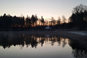 Une scène paisible au bord d’un lac, avec une structure en forme de tente au centre, entourée d’arbres et d’une surface d’eau calme qui reflète le ciel.
