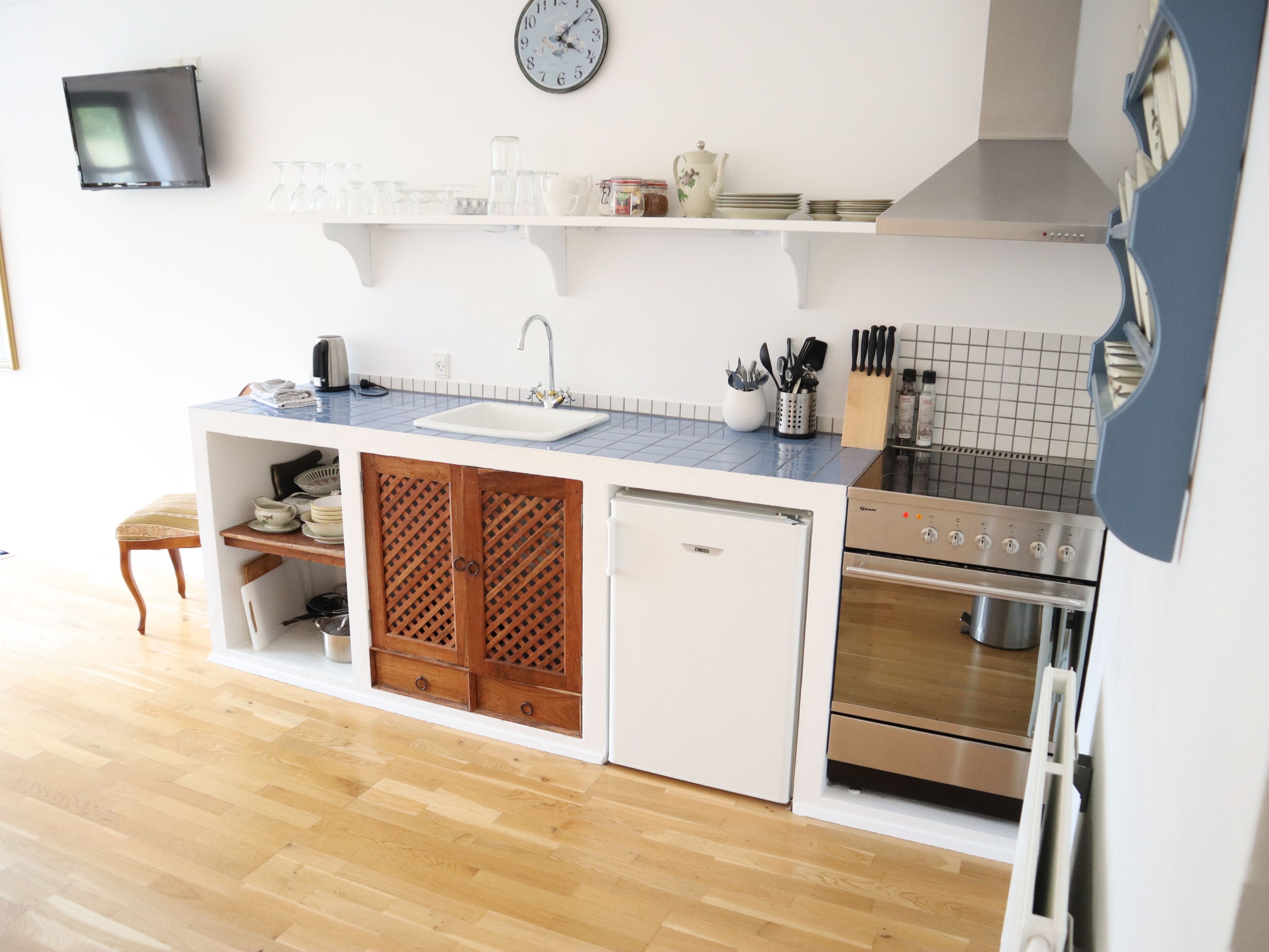 A compact kitchen with a sink, stove, and refrigerator, featuring a wooden cabinet and a clock on the wall.