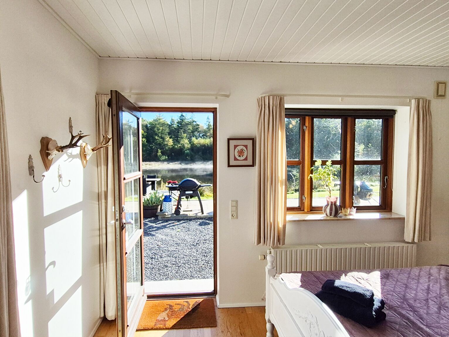 A bedroom with a bed, a window with curtains, a radiator, and a door leading to an outdoor area with a table and chairs.