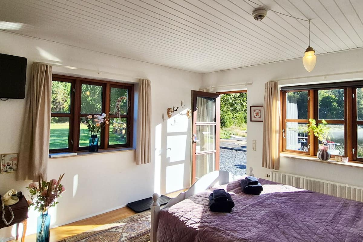 Ein Schlafzimmer mit einem großen Bett, einem Fenster mit Vorhängen, einer Tür, die nach draußen führt, und einem kleinen Tisch mit einem Blumenstrauß.
