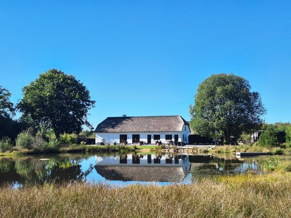 Un bâtiment blanc avec un toit de chaume, entouré d’arbres et bordé par une étendue d’eau calme.