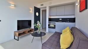 A modern living room with a flat-screen TV, a wooden coffee table, a vase with flowers, a sofa with a yellow pillow, and a kitchen area with grey cabinets and a black countertop. - 2-room apartment panoramic sea view (Rayol-Canadel-sur-Mer)