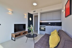 Ein modernes Wohnzimmer mit Flachbildfernseher, Couchtisch aus Holz, einer Vase mit Blumen, einem Sofa mit gelbem Kissen und einer Küchenzeile mit grauen Schränken und einer schwarzen Arbeitsplatte.