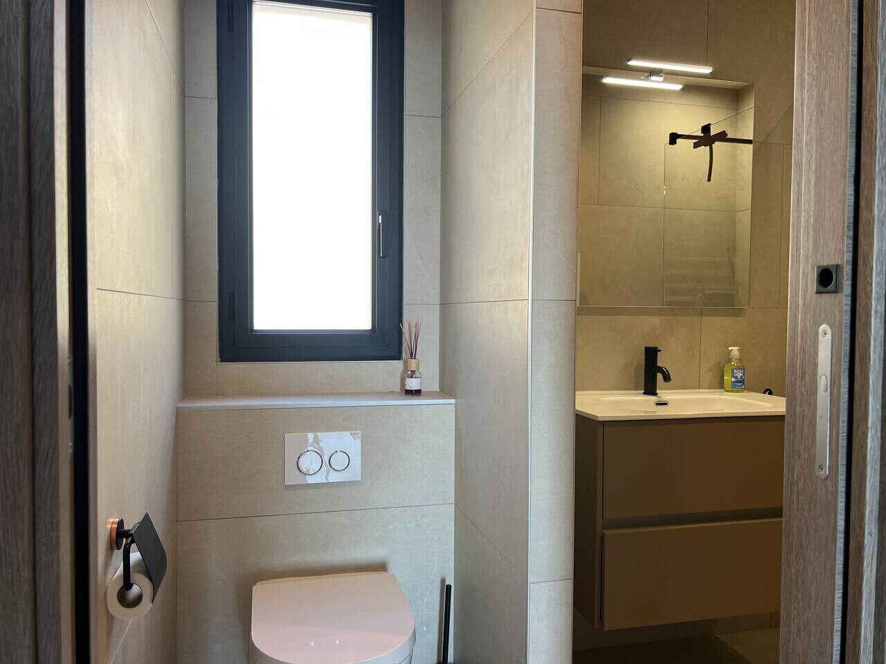 Een moderne badkamer met toilet, wastafel en een raam boven de wastafel.