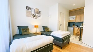 A room with two beds, a small table with a lamp, and a kitchenette in the background.